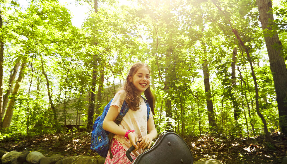 A young girl with long brown hair holding a violin case outside at Usdan.