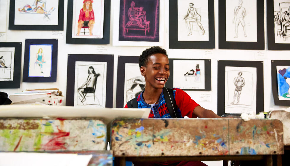 Young boy sitting at an art studio desk smiling with paintings pinned up behind him.