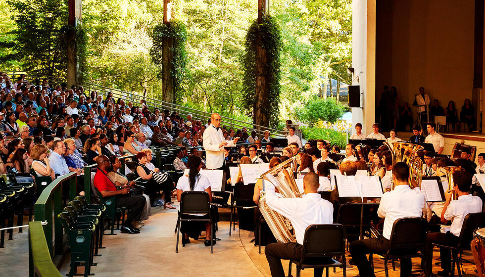 Conductor in front of band in Amphitheater surrounded by green nature.