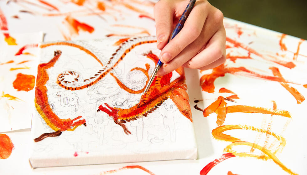 Closeup of child's hand painting an orange dragon on canvas.