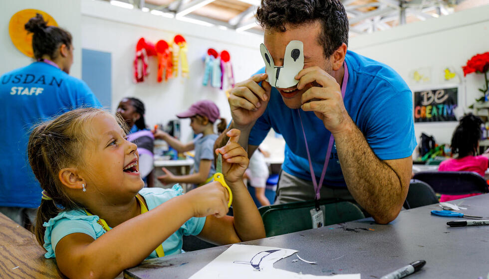 Image of a small girl in a light blue shirt holding yellow scissors smiling at a camp counselor holding a mask.