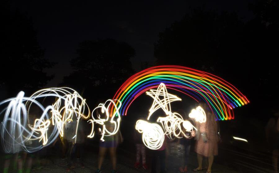 Children in the dark using lights to make shapes with light and motion. We see a rainbow and a star and squiggles.