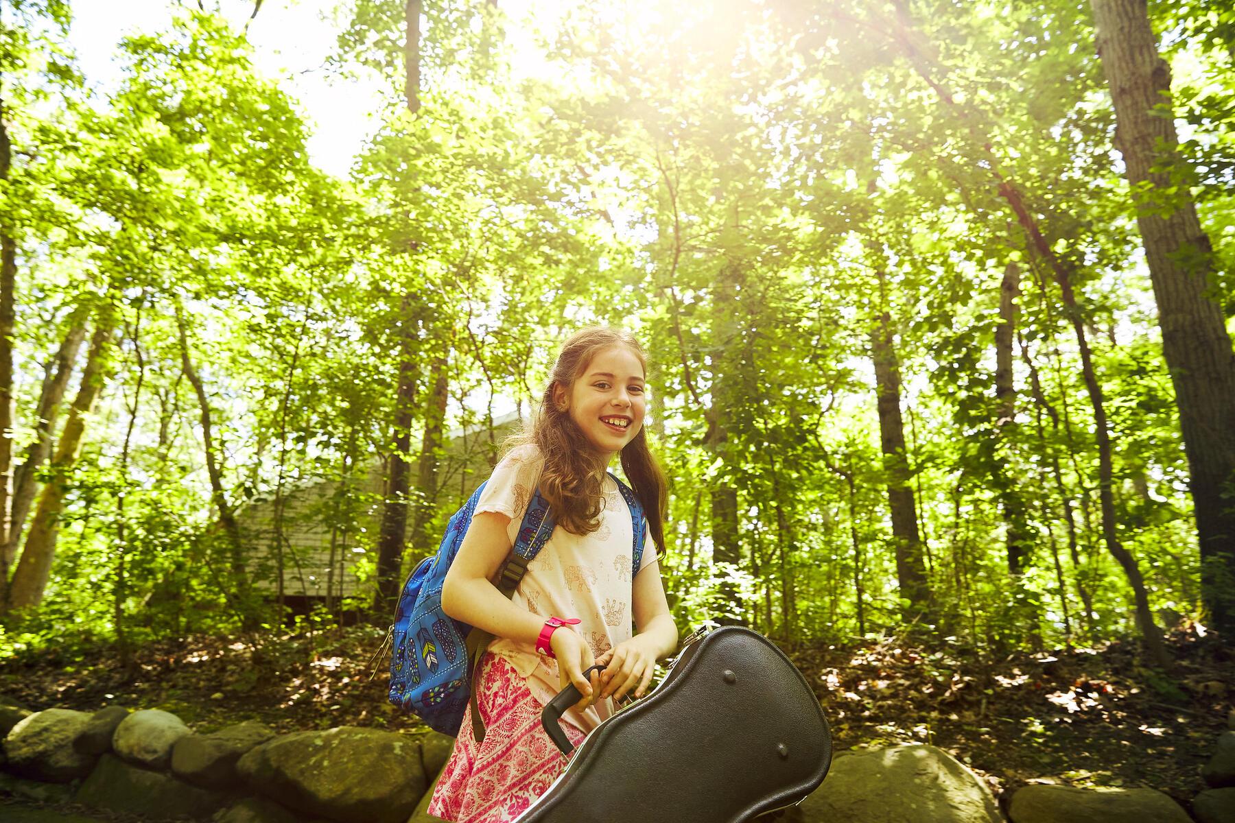 A young girl with long brown hair holding a violin case outside at Usdan