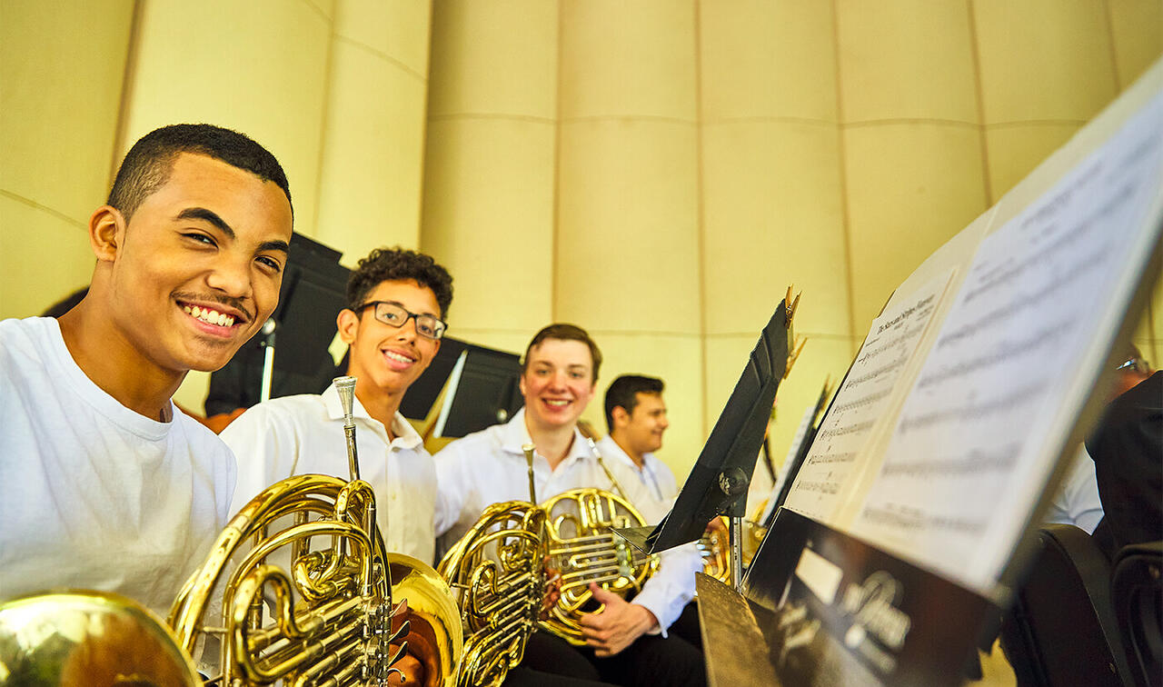 Three smiling students in white shirts holding french horns in front of music stands.