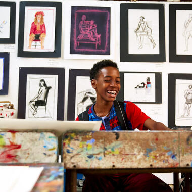 Young boy sitting at an art studio desk smiling with paintings pinned up behind him.