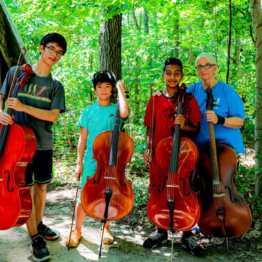 Cellists posing with their cellos in the woods.