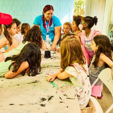 Students standing around an art table listening to their teacher Students standing around an art table listening to their teacher