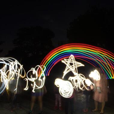 Children in the dark using lights to make shapes with light and motion. We see a rainbow and a star and squiggles.